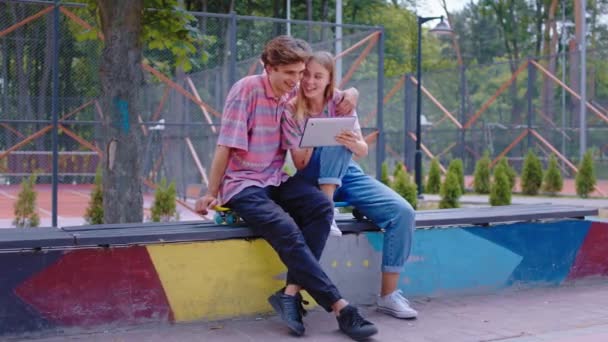 Charismatic casal bonito em um parque de skate moderno sentado nas cadeiras passar o tempo juntos fora eles usando um gadget tablet eletrônico — Vídeo de Stock