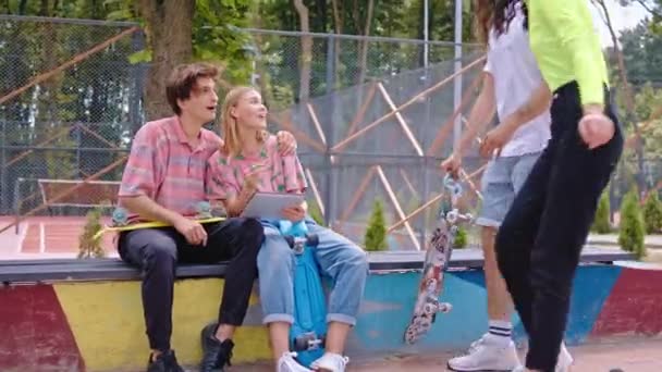 Молода пара зустрічає деяких друзів на вулиці в сучасному скейт-парку, а їхнє перебування на електронному планшеті щасливе вони спілкуються разом — стокове відео
