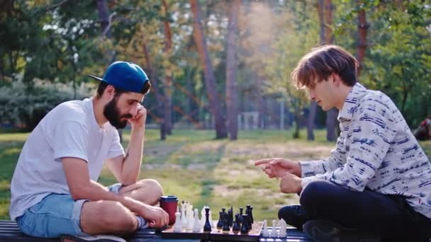 Mitten im Park sitzen zwei Teenager auf dem Parkstuhl und spielen ein Schachspiel, das sie konzentriert miteinander verbringen — Stockvideo