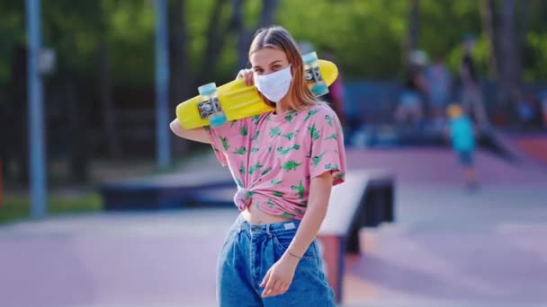 Mit Schutzmaske und blonden Haaren hält eine Dame ihr Skateboard in einem städtischen Skatepark am Konzept des Coronavirus 2019 fest — Stockvideo