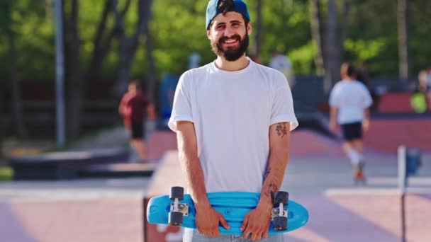 Vysoký chlap s velkým úsměvem před kamerou v moderním skate parku drží svůj skateboard před zahájením jízdy — Stock video