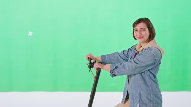 Χαρισματικό χαμόγελο χαριτωμένο κυρία με κοντά μαλλιά σε ένα πράσινο στούντιο τοίχο ποζάρουν μπροστά από την κάμερα, ενώ κρατώντας ηλεκτρικό σκούτερ της — Αρχείο Βίντεο