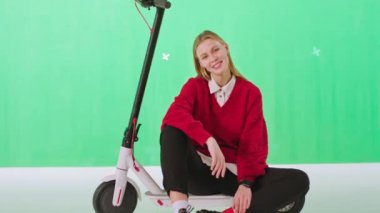 Stüdyonun yeşil arka planında kameranın önünde elektrikli scooterının üzerinde otururken büyük bir gülümsemeyle poz veren güzel bayan kavramı. 4k