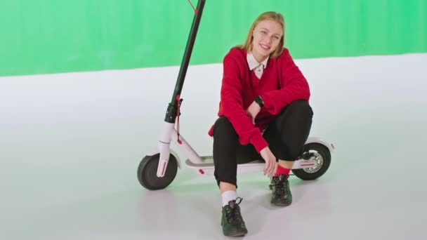 Büyük yeşil stüdyo sarı saçlı kadın elektrikli scooterın üzerinde otururken doğrudan kameraya bakıyor. 4k — Stok video