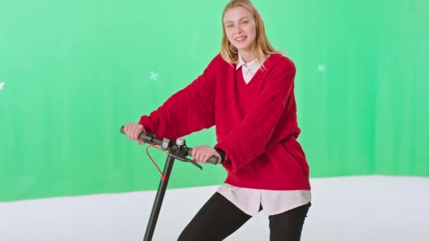 Kameranın önünde, yeşil bir stüdyoda modern elektrikli scooterını tutarken sarı saçlı harika bir kadın pozu veriyor. — Stok video