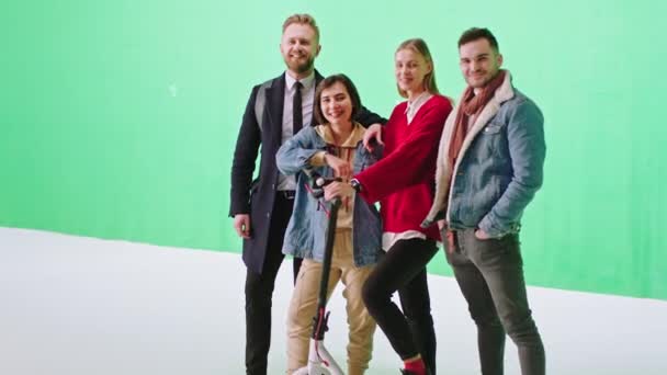 Carismático grupo de amigos en un estudio de fondo verde posando delante de la cámara y sonriendo a lo grande sosteniendo un scooter eléctrico — Vídeos de Stock