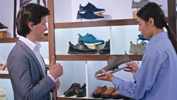 Primer plano de la cámara hombre cliente en un traje llegó a la zapatería para comprar algunos zapatos que discute con el asiático señora consultora — Vídeos de Stock