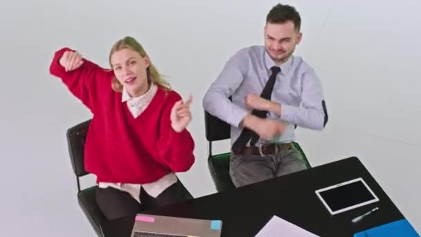 Voor de camera jonge vrouw en man zitten op hun werkplek en dansen gelukkig kijken recht naar de camera — Stockvideo