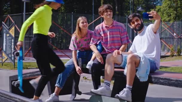 Attraente gruppo di amici hipster giovani e carismatici signore e ragazzi che scattano selfie in un moderno skate park si siedono sulla sedia del parco e in posa per un selfie hanno un grande sorriso — Video Stock