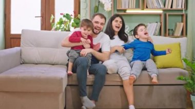 Kanepede büyük bir gülümsemeyle mutlu bir aile zamanı. İki çocuk ve ebeveynler televizyonda bir şey izliyorlar. Gülüyorlar ve kameranın önünde eğleniyorlar.