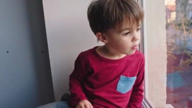 Evde karantinada oturan sevimli küçük çocuk, salgın bittikten sonra dışarı çıkmayı bekleyen üzgün bir suratla pencereden bakıyor. Yeni Covid-19. 4k