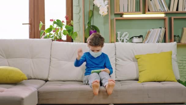 Glücklicher kleiner Junge 5 Jahre alt mit dem Smartphone ein Spiel zu spielen, während zu Hause bleiben in Quarantäne und trägt Schutzmaske Konzept des Coronavirus 2019 — Stockvideo