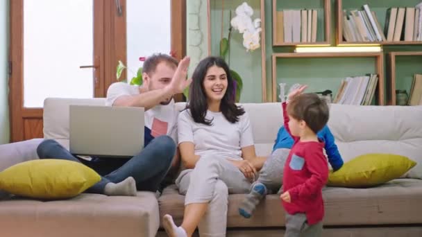 Keluarga muda yang bahagia dengan wajah tersenyum menghabiskan waktu bersama-sama sambil duduk di sofa mereka memeluk dan bermain dengan satu sama lain — Stok Video