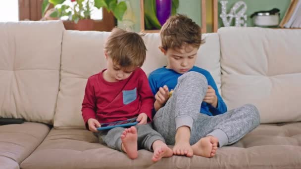 Μπροστά από την κάμερα στον καναπέ δύο μικρά παιδιά αδέλφια παίζουν στο smartphone περνούν το χρόνο τους μαζί — Αρχείο Βίντεο