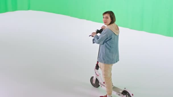 Σε ένα μεγάλο πράσινο φόντο κυρία στούντιο στέκεται μπροστά από την κάμερα, ενώ κρατά ηλεκτρικό σκούτερ της — Αρχείο Βίντεο