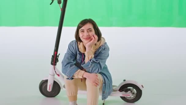 Piękna młoda kobieta siedząca na skuterze elektrycznym patrząc przed kamerę w dużym zielonym studio tła. 4k — Wideo stockowe