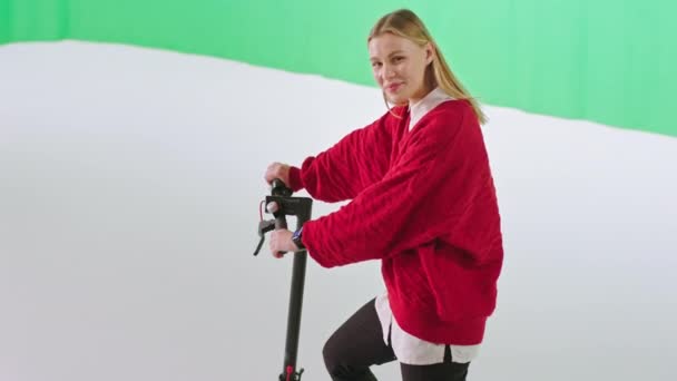 Genç ve karizmatik bir kadın elektrikli scooterını tutuyor ve yeşil duvar stüdyosu olan geniş bir arkaplanda kameraya bakıyor. — Stok video