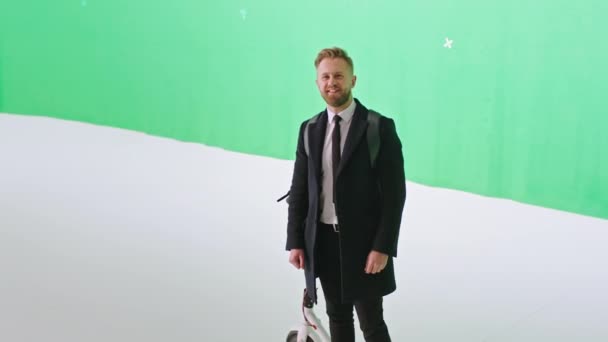 Ένας άντρας με μαύρο κοστούμι, υπάλληλος γραφείου, ποζάρει μπροστά στην κάμερα, σε ένα μεγάλο πράσινο στούντιο, κρατώντας το ηλεκτρικό του σκούτερ. — Αρχείο Βίντεο