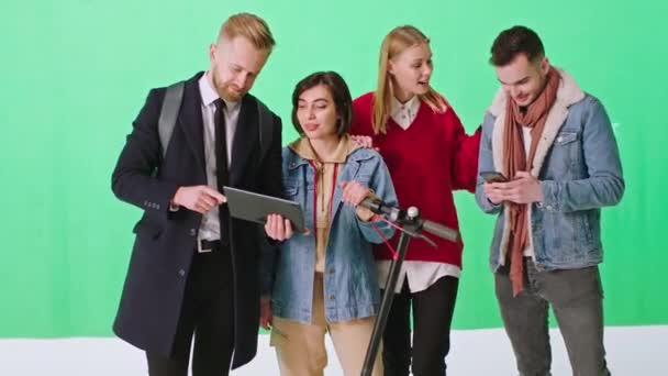 Beau groupe de jeunes gens socialisant ensemble ils utilisent une tablette électronique moderne et scooter électrique dans un grand studio de fond vert — Video