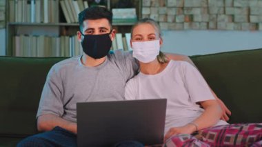Mutlu ve karizmatik bir çift karantinada kanepede koruyucu bir maske takıp doğrudan kameraya bakıyor.