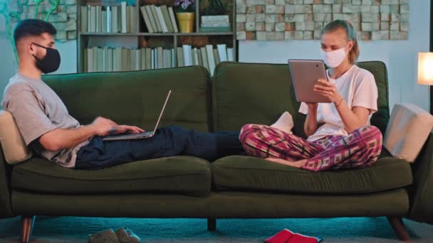 家からオンラインで勉強ソファの上に保護マスクを持つ良い探している女性と男はコロナウイルスのパンデミックです — ストック動画