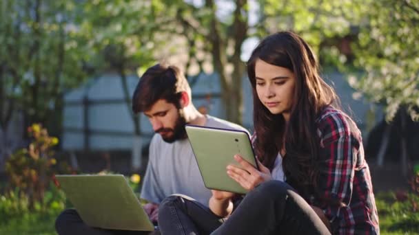 Charismatische vrouw en man tijdens het zitten op het gras ze geconcentreerd werken of studeren online met behulp van de gadgets in een per zonnige dag — Stockvideo