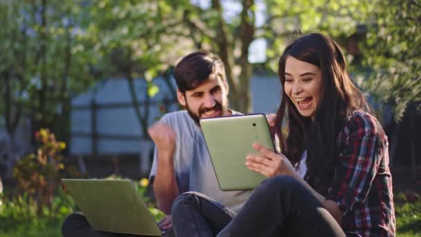 En la hierba sonriendo linda hembra y su amigo chico guapo juntos estudiando al aire libre se divierten juntos usando el portátil y la tableta para trabajar — Vídeo de stock