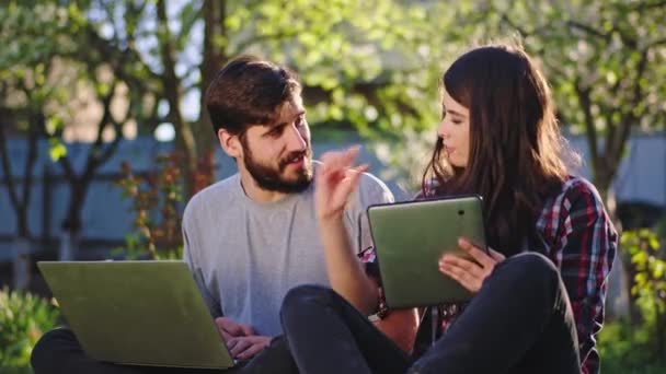 En el jardín pareja guapa tener una conversación juntos señora ayudar al hombre cómo terminar su proyecto de la universidad utilizando un ordenador portátil y tableta. Disparo en ARRI Alexa Mini — Vídeo de stock