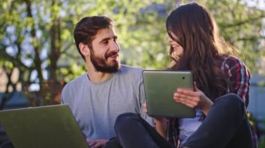 Çekici genç bir kadın ve yakışıklı bir erkek, dışarıda çimlerin üzerinde oturup sohbet ediyorlar ve tablet ve dizüstü bilgisayarlarını kullanarak internet üzerinden çalışıyorlar.