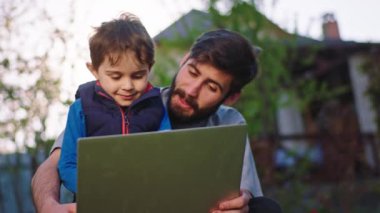 Kameranın önünde yakışıklı ve mutlu çocuk çimlerin üzerinde oturup babasının dizüstü bilgisayarıyla oynarken genç babasıyla vakit geçirdiği için çok heyecanlı.