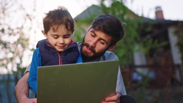 Na frente da câmera bonito e feliz menino muito animado que ele passa o tempo com seu jovem pai enquanto se senta na grama e jogando no laptop pais — Vídeo de Stock