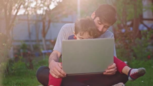 Στον κήπο ελκυστικό μικρό αγόρι και ο μπαμπάς του έχουν μια καλή στιγμή μαζί παίζουν ένα παιχνίδι, ενώ χρησιμοποιώντας το laptop απολαμβάνοντας το χρόνο μαζί. Πυροβολήθηκε στο ARRI Alexa Mini — Αρχείο Βίντεο