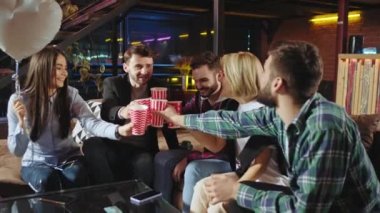 Modern stil bir dairede oturma odasında genç bir grup arkadaş renkli bardaklarla kutlama yapıyor ve birlikte vakit geçiriyorlar.