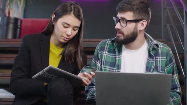 One charismatic guy and Japanese smiling large lady in a modern office center while sitting on the office stairs doing a order online using a credit card and electronic tablet — Stock Video