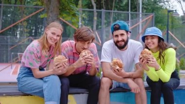 Modern bir kaykay parkında çok ırklı arkadaşlardan oluşan bir kamera grubunun önünde biraz fast food burgeri alıp kameranın önünde yemek yiyorlar.