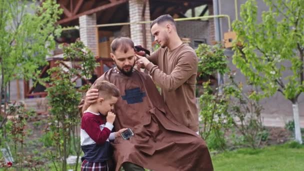 Atraktivní otec a jeho syn sedí na zahradě vedle malého chlapce a hrají si na smartphonu, zatímco jeho táta se stříhá — Stock video