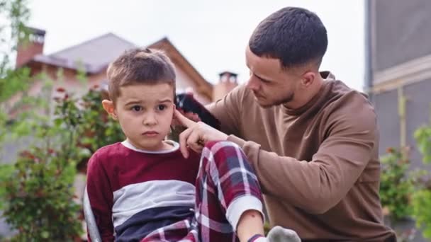 In een zonnige dag in de tuin thuis maakt de professionele kapper een knipbeurt voor een schattig grappig jongetje — Stockvideo