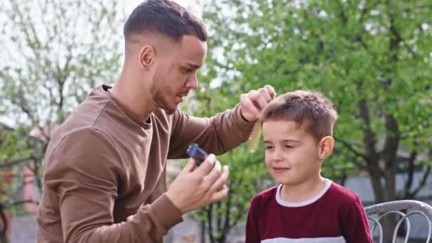 Par une journée ensoleillée dans le jardin un petit garçon son grand frère faire une coupe de cheveux avec une tondeuse à cheveux pendant Coronavirus en quarantaine — Video