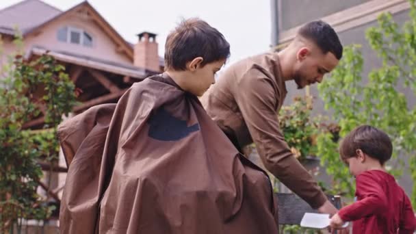 В саду дома харизматичный мальчик готовится к стрижке парикмахер готовит электрический клипер другой маленький мальчик взять щетку — стоковое видео
