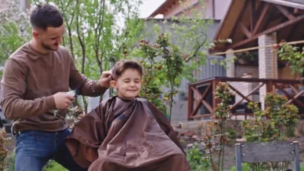 Muy divertido lindo niño pequeño tiene un corte de pelo en el jardín que hacen una cara linda delante de la cámara barbero hombre concentrado trabajando con un cortador electrónico — Vídeos de Stock