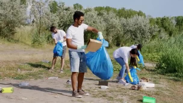 Volontärer som plockar upp soporna mitt i naturen håller ihop stora blå plastpåsar i skyddshandskar och städar upp skräp — Stockvideo