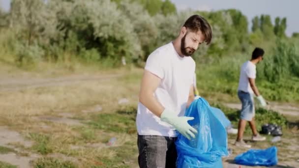 Delante de la cámara en la playa junto al chico del lago voluntario con una gran bolsa de plástico mirando directamente a la cámara mientras él y sus amigos recogen la basura — Vídeo de stock