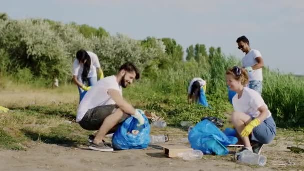 На пляжі біля озера група багаторасових добровольців разом працюють над пляжем, збираючи сміття на блакитних пластикових пакетах — стокове відео