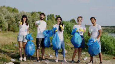 Kamera önünde çok ırklı gönüllüler mavi plastik poşetlerle poz veriyor ve plastik poşetleri topladıktan sonra büyük gösteriyor. 4k