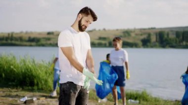 Çöpleri geri dönüştürme kavramı. Koruyucu eldiven giyen gönüllü genç adam plajdaki çöpleri temizlemeye başlıyor. Diğer gönüllüler çöpleri temizliyor.