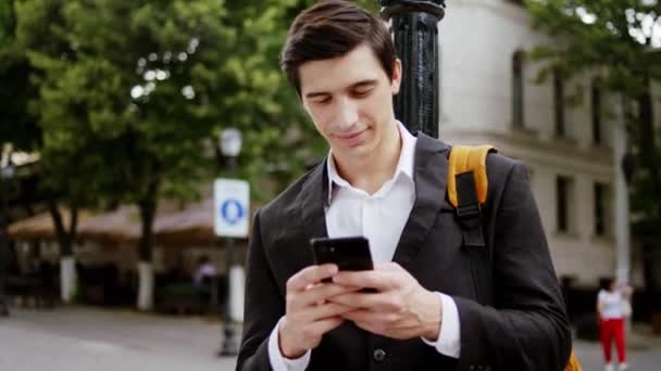 Concentrado con una bonita sonrisa joven de negocios escribiendo algo en su teléfono inteligente en medio de la calle. Disparo en ARRI Alexa Mini — Vídeo de stock