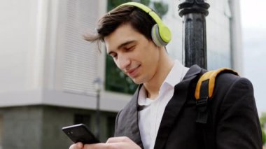 Siyah takım elbiseli genç adam sokağın ortasında kameraya yakın duruyor kulaklıkla müzik dinliyor ve akıllı telefonuyla bir şeyler izliyor.
