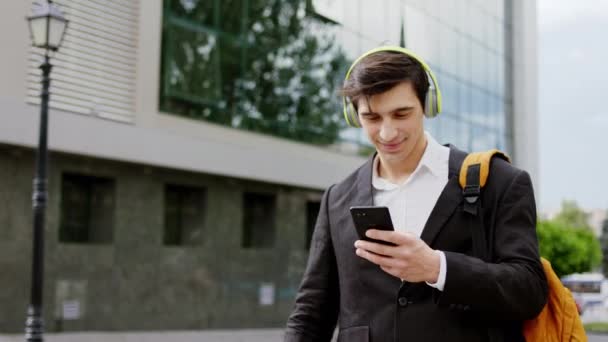 Hombre atractivo en un traje negro tomar su teléfono inteligente viendo algo que sonríe grande escuchar música en los auriculares — Vídeo de stock