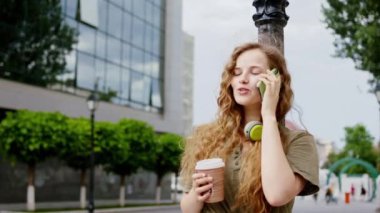 Caddenin ortasında elinde bir fincan kahve tutarken mutlu ve gülümseyen çekici bir kadın akıllı telefonuyla tartışıyor. ARRI Alexa Mini 'de çekilen