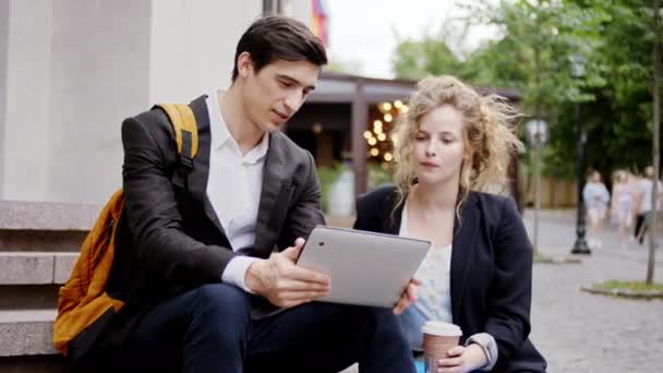 In the middle of the street charismatic and attractive businessman and his colleague blonde hair lady stopping on some steps take a look on the electric tablet to analyzing something. Shot on ARRI — Stock Video